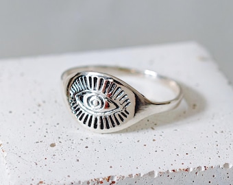 Può includere: Un anello d'argento con una faccia ovale raffigurante un occhio. Il design dell'occhio presenta linee dettagliate e una pupilla centrale. L'anello è esposto su una superficie bianca strutturata, evidenziando il design e l'artigianato.