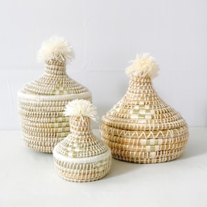 Könnte beinhalten: Drei geflochtene Körbe mit weißen Bommeln oben. Die Körbe sind in verschiedenen Größen und aus natürlichen Materialien gefertigt.