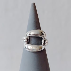 Könnte beinhalten: Ein glänzender Silberring mit einem einzigartigen Design. Der Ring hat ein dickes, abgerundetes Band, das eine rechteckige Form mit abgerundeten Ecken bildet. Er wird auf einem grauen, kegelförmigen Ständer präsentiert.