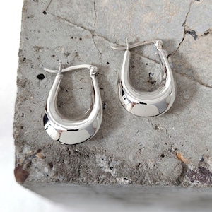 May include: A pair of polished silver hoop earrings. The earrings have a rounded, U-shaped design with a smooth, reflective surface. They are displayed on a textured, gray concrete surface, showcasing their bright, metallic finish.