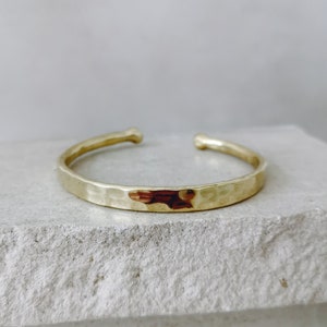 Könnte beinhalten: Ein goldfarbenes Armband in Manschettenform mit einer gehämmerten, strukturierten Oberfläche. Das Armband hat ein offenes Design mit abgerundeten, glatten Enden und eine reflektierende Oberfläche, die das Licht auf der Struktur einfängt.