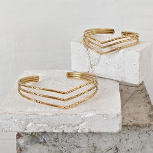 Op de afbeelding: Twee goudkleurige, open manchetarmbanden met een getextureerde afwerking. Elke armband heeft een chevron-ontwerp met drie parallelle banden. De armbanden worden getoond op een wit en grijs getextureerd oppervlak, met een effen witte achtergrond.