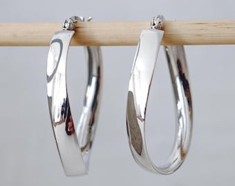 May include: A pair of polished silver hoop earrings. The earrings are a flattened oval shape, with a smooth, reflective surface. They are suspended from a wooden dowel against a light gray background. The earrings have a simple, modern design.