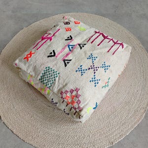 Peut inclure: Un coussin de sol carré en tissu blanc cassé orné de motifs géométriques brodés dans des couleurs vives, notamment rose vif, vert néon, orange, bleu et noir. Le coussin présente une surface texturée avec divers motifs au point de croix.