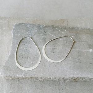 Könnte beinhalten: Ein Paar silberner, tropfenförmiger Creolen. Die Ohrringe haben eine glatte, polierte Oberfläche und ein minimalistisches Design. Sie werden auf einer strukturierten, grauen Steinoberfläche präsentiert.