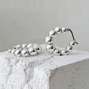 May include: A pair of silver hoop earrings with a beaded design. Each earring is composed of multiple small, shiny, spherical beads. One earring is open, while the other is closed. The earrings are displayed on a white, textured surface.