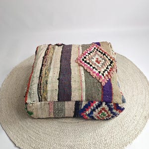 Könnte beinhalten: Ein quadratisches, mehrfarbiges Bodenkissen mit Patchwork-Design. Das Kissen hat vertikale Streifen in verschiedenen Farben, darunter Beige, Lila und Grün. Ein dekorativer, rautenförmiger Aufnäher ist oben aufgenäht. Das Kissen liegt auf einem runden, gewebten Teppich.