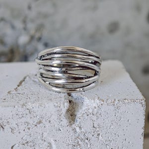 Peut inclure: Une bague argentée avec un motif tissé. La bague présente plusieurs bandes de métal entrelacées, créant un aspect texturé et superposé. La bague est présentée sur une surface blanche et texturée.