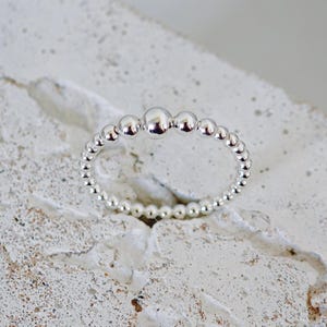 Könnte beinhalten: Ein Silberring, der aus einem durchgehenden Band kleiner, polierter, kugelförmiger Perlen besteht. Das Design zeigt eine einzelne, etwas größere Mittelperle, die von zwei kleineren Perlen flankiert wird, während das restliche Band aus gleichmäßigen Silberkugeln besteht.