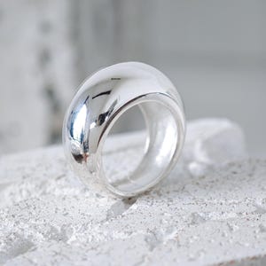 Könnte beinhalten: Ein polierter silberfarbener Ring mit einer dicken, abgerundeten, kuppelförmigen Ringschiene. Die Oberfläche des Rings ist hochglänzend, weist klare Lichtreflexe auf und hat ein glattes, geschwungenes Profil, das eine solide Kreisform bildet.