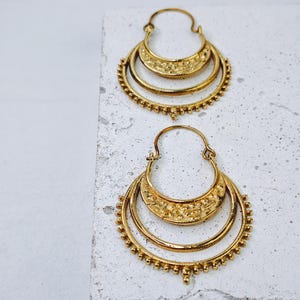 Può includere: Due orecchini a cerchio color oro. Ogni orecchino presenta una forma a mezzaluna con tre livelli. Il livello superiore ha un design strutturato, mentre il livello inferiore ha piccoli abbellimenti rotondi. Gli orecchini sono esposti su una superficie bianca.