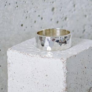 Könnte beinhalten: Ein breiter Silberring mit einer gehämmerten, strukturierten Oberfläche, die ein reflektierendes, facettiertes Aussehen erzeugt. Das Metall hat ein helles, poliertes Finish, das das Licht auf seiner unregelmäßigen Oberfläche einfängt.