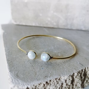 Può includere: Un bracciale aperto color oro con due pietre rotonde di colore chiaro. Il bracciale è posto su uno sfondo neutro, evidenziando il design e i materiali del gioiello. Le pietre sembrano essere pietre di luna.