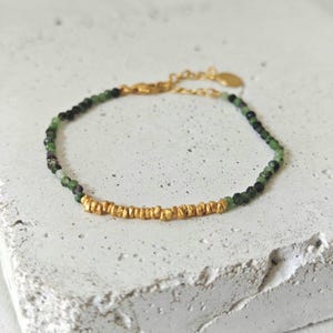 Op de afbeelding: Een delicate gouden armband met een rij gouden kralen en een streng kleine groene en zwarte kralen. De armband wordt gepresenteerd op een wit oppervlak, wat het elegante ontwerp en het kleurcontrast benadrukt.