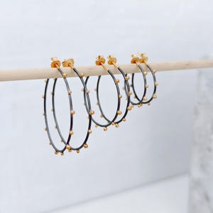 May include: A set of four hoop earrings. The earrings have a dark gray band with small gold-colored beads evenly spaced around the hoop. The posts and the beads are gold-colored. The earrings are displayed on a wooden dowel against a white background.