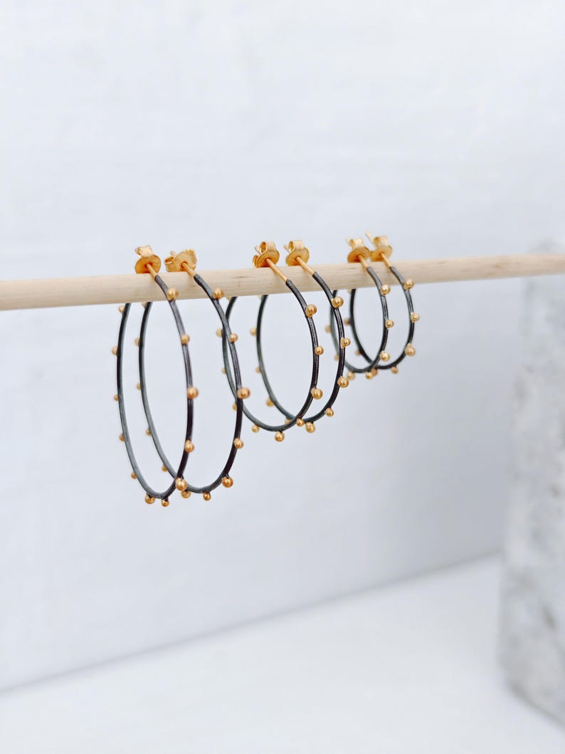 May include: A set of four hoop earrings. The earrings have a dark gray band with small gold-colored beads evenly spaced around the hoop. The posts and the beads are gold-colored. The earrings are displayed on a wooden dowel against a white background.