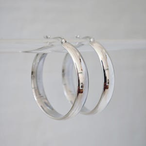May include: A pair of shiny, silver hoop earrings. The earrings are round and have a smooth, reflective surface. They are suspended against a light background, highlighting their polished appearance. The hoops are a classic jewelry design.