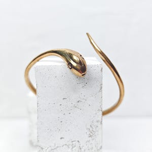May include: A gold-colored snake-shaped cuff bracelet. The bracelet features a detailed snake head with small black eyes. The body of the snake curves around, creating an open cuff design. The bracelet is displayed on a white, textured surface.