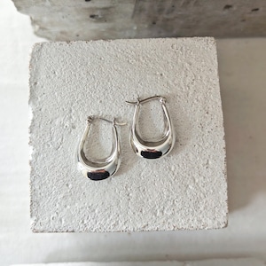 May include: A pair of silver hoop earrings with a teardrop shape. The earrings are highly polished and reflect light, creating a shiny appearance. They are displayed on a textured, light-colored surface.
