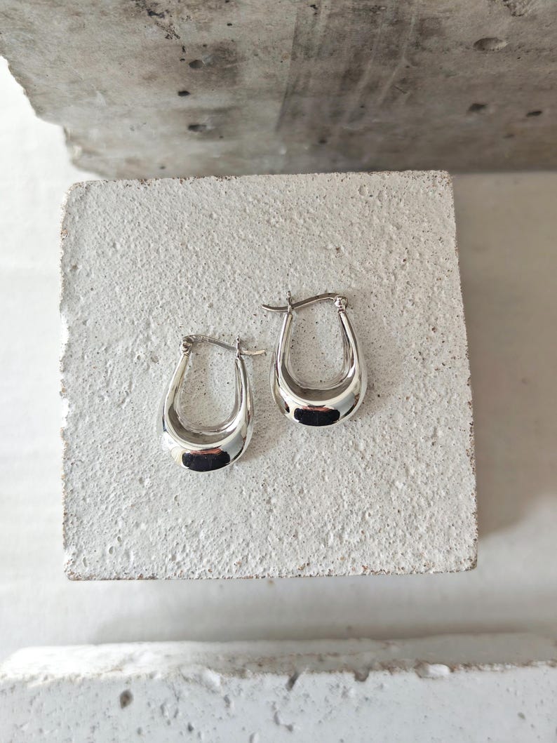 May include: A pair of silver hoop earrings with a teardrop shape. The earrings are highly polished and reflect light, creating a shiny appearance. They are displayed on a textured, light-colored surface.