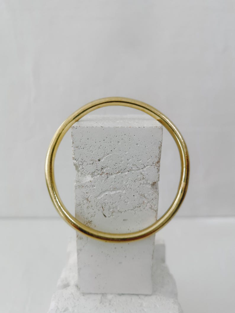 May include: A gold-colored bangle bracelet. The circular bracelet is displayed against a white, textured background. The bangle is a simple, elegant design, suitable for everyday wear.