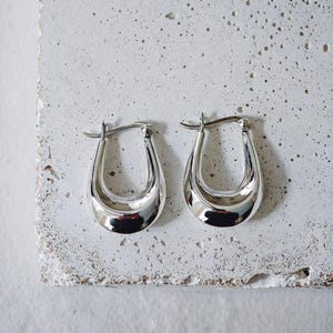 May include: A pair of polished silver hoop earrings. The earrings have a smooth, curved design with a shiny, reflective surface. The earrings are displayed on a textured, light gray surface.