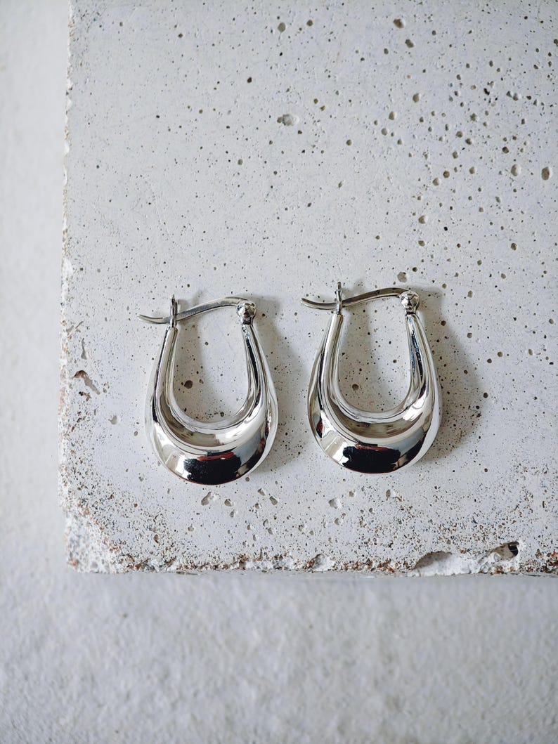 May include: A pair of polished silver hoop earrings. The earrings have a smooth, curved design with a shiny, reflective surface. The earrings are displayed on a textured, light gray surface.