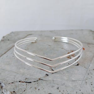 Op de afbeelding: Een zilverkleurige manchetarmband met een chevron-ontwerp. De armband heeft drie parallelle banden die in het midden een V-vorm vormen. Het open ontwerp maakt het gemakkelijk om te dragen. De armband wordt op een grijze ondergrond getoond.