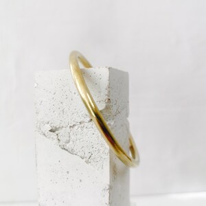 May include: A gold-colored bangle bracelet. The smooth, rounded bracelet is displayed on a textured, white, rectangular block. The background is a plain white.