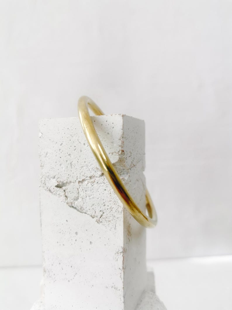 May include: A gold-colored bangle bracelet. The smooth, rounded bracelet is displayed on a textured, white, rectangular block. The background is a plain white.