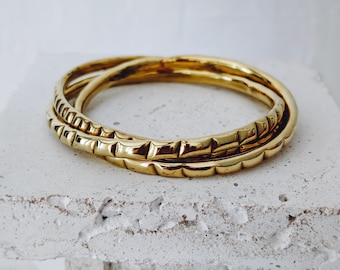 Puede incluir: Tres pulseras doradas, dos lisas y una con un patrón rectangular texturizado. Las pulseras están apiladas sobre una superficie blanca texturizada, creando una exhibición simple pero elegante. La joyería está hecha de un material metálico brillante.