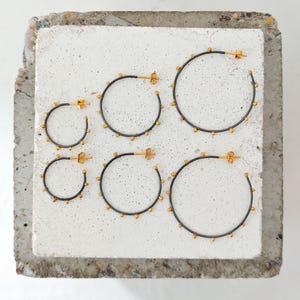 May include: Six pairs of hoop earrings in varying sizes. The earrings have a dark metal finish with small gold-colored beads and gold-colored posts. The earrings are displayed on a light gray, textured surface.
