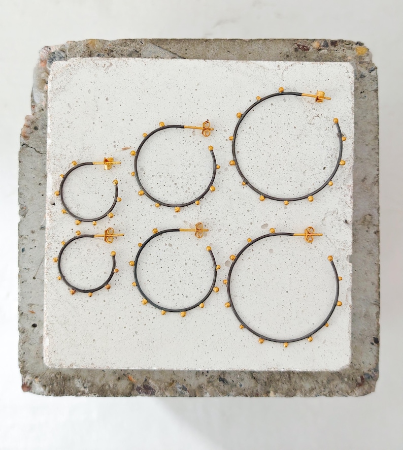 May include: Six pairs of hoop earrings in varying sizes. The earrings have a dark metal finish with small gold-colored beads and gold-colored posts. The earrings are displayed on a light gray, textured surface.