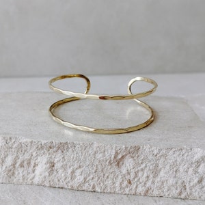 Pode incluir: Um bracelete de metal martelado dourado. O bracelete é aberto e tem um design simples e minimalista.
