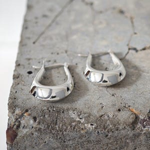 May include: A pair of shiny silver hoop earrings. The earrings have a rounded, teardrop shape and a polished finish. They are displayed on a rough, gray concrete surface.