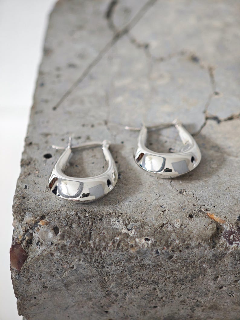 May include: A pair of shiny silver hoop earrings. The earrings have a rounded, teardrop shape and a polished finish. They are displayed on a rough, gray concrete surface.