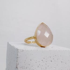 Könnte beinhalten: Ein goldfarbener Ring mit einem großen, tropfenförmigen, facettierten, hellrosa Edelstein. Der Edelstein ist in einer goldenen Lünette gefasst und auf einem zarten Goldband montiert. Der Ring wird auf einer weißen, strukturierten Oberfläche präsentiert.