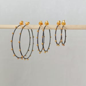 May include: Three pairs of hoop earrings in varying sizes. The hoops are dark gray with small, evenly spaced gold-colored beads. The earring posts and the beads are gold-colored. The earrings are displayed on a wooden dowel against a light background.