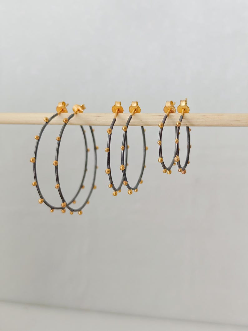 May include: Three pairs of hoop earrings in varying sizes. The hoops are dark gray with small, evenly spaced gold-colored beads. The earring posts and the beads are gold-colored. The earrings are displayed on a wooden dowel against a light background.