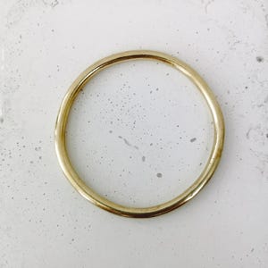 May include: A gold-colored, circular bangle bracelet. The bracelet is a simple, minimalist design with a smooth, polished surface. The bangle is set against a speckled, light-colored background.