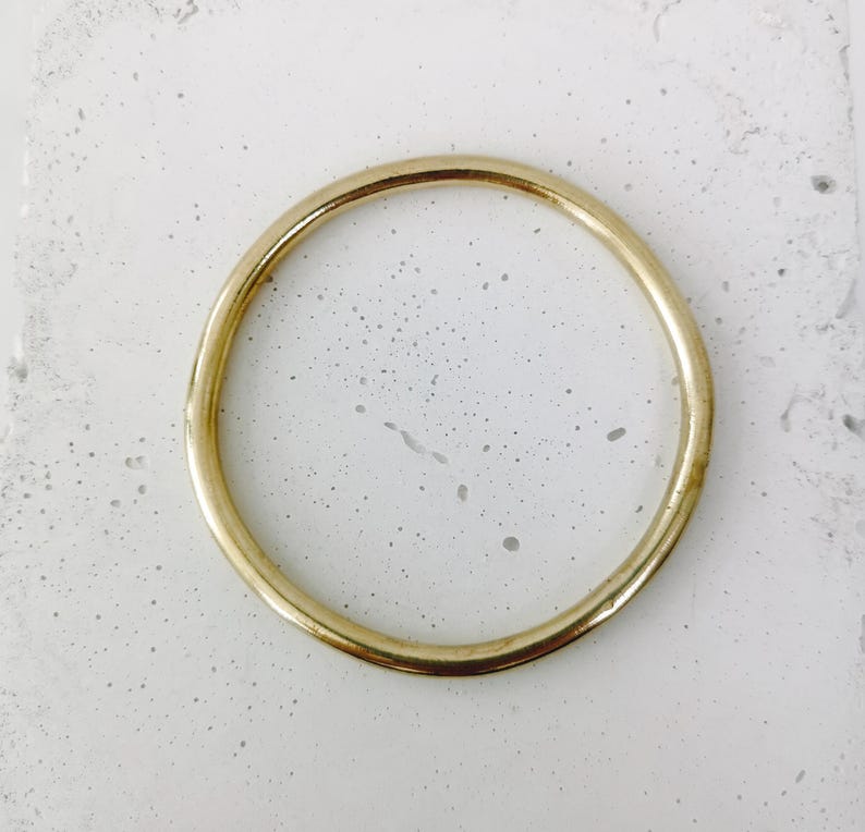 May include: A gold-colored, circular bangle bracelet. The bracelet is a simple, minimalist design with a smooth, polished surface. The bangle is set against a speckled, light-colored background.