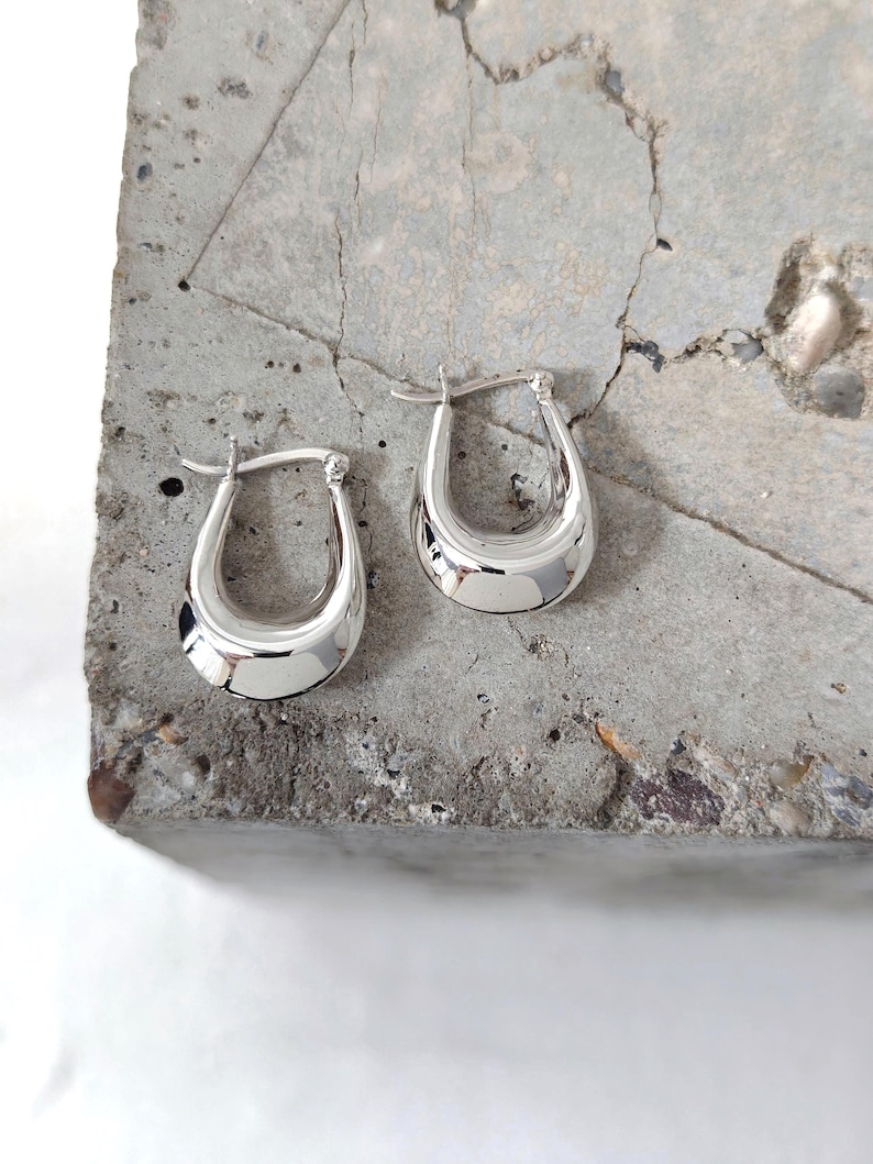 May include: A pair of polished silver hoop earrings. The earrings have a rounded, U-shaped design with a smooth, reflective surface. They are displayed on a textured, gray concrete surface, showcasing their bright, metallic finish.