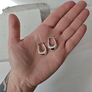 May include: Two silver-colored, small hoop earrings. The earrings have a rounded, teardrop shape. The earrings are smooth and shiny, reflecting light. The earrings are displayed on a light-colored background.