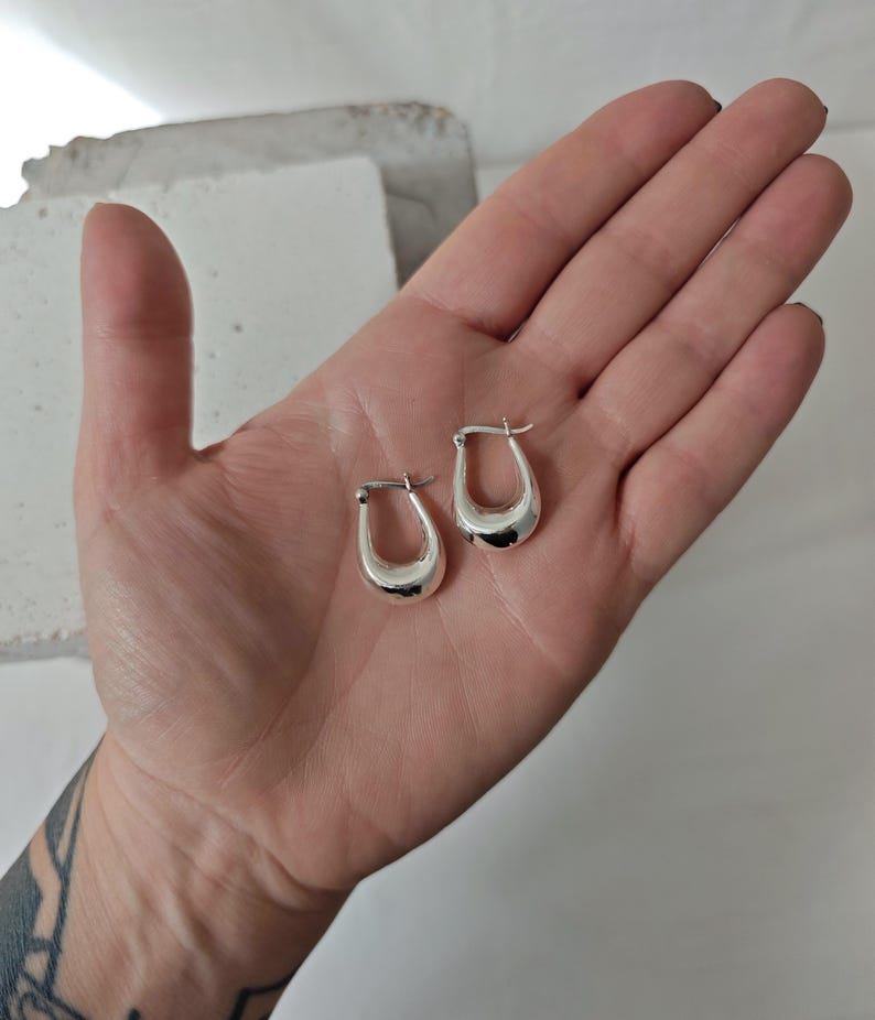 May include: Two silver-colored, small hoop earrings. The earrings have a rounded, teardrop shape. The earrings are smooth and shiny, reflecting light. The earrings are displayed on a light-colored background.