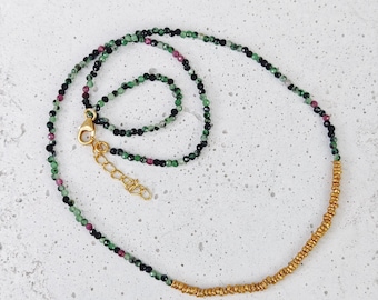 May include: A delicate necklace featuring a strand of small, faceted beads in shades of green, black, and pink. The necklace transitions to a section of small, gold-colored beads and is finished with a gold-toned clasp.