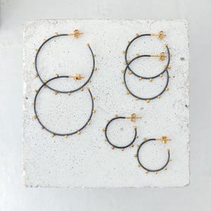 May include: Several pairs of hoop earrings in varying sizes. The earrings have a dark gray finish with small gold-colored studs around the hoops. The posts and the inner part of the hoops are gold-colored. The earrings are displayed on a light gray surface.
