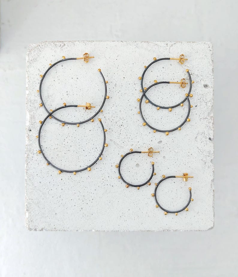 May include: Several pairs of hoop earrings in varying sizes. The earrings have a dark gray finish with small gold-colored studs around the hoops. The posts and the inner part of the hoops are gold-colored. The earrings are displayed on a light gray surface.
