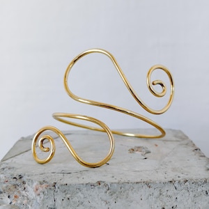 Puede incluir: Un brazalete dorado con un diseño en espiral. El brazalete se envuelve alrededor del brazo con dos espirales. La joya está hecha de un alambre de metal fino y continuo y descansa sobre una superficie gris texturizada.