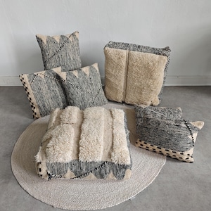 May include: A collection of six square and rectangular throw pillows featuring a mix of black and white woven fabric and cream-colored shaggy wool panels. The pillows display geometric patterns, braided trim details, and textured surfaces.