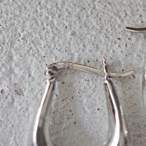 May include: Close-up of a pair of silver hoop earrings. The earrings have a smooth, polished finish and a teardrop shape. The clasp is visible, and the metal is stamped with "925". The background is a textured, off-white surface.